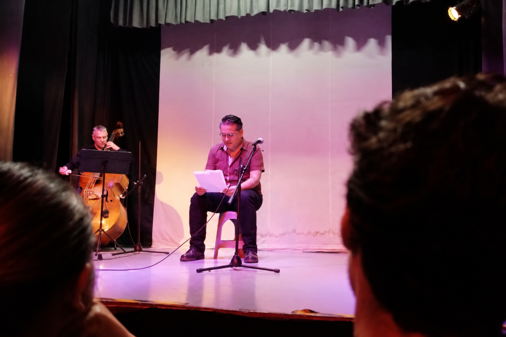 Participante realiza una lectura en el escenario acompañado por Miguel Trejo al contrabajo durante la Tardeada Surrealista del Poeta Muerto.